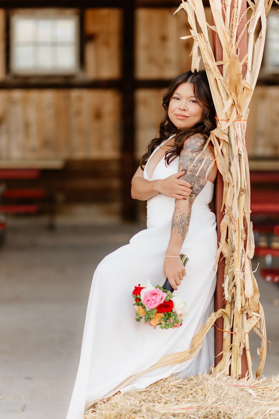 Bride in white gown holding colorful bouquet sits on hay bale beside dried corn stalks in rustic barn.