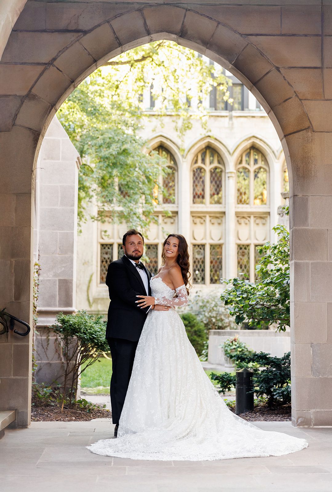 Bride and groom posing under a stone arch at a Gothic-style venue, bride in lace off-shoulder gown.