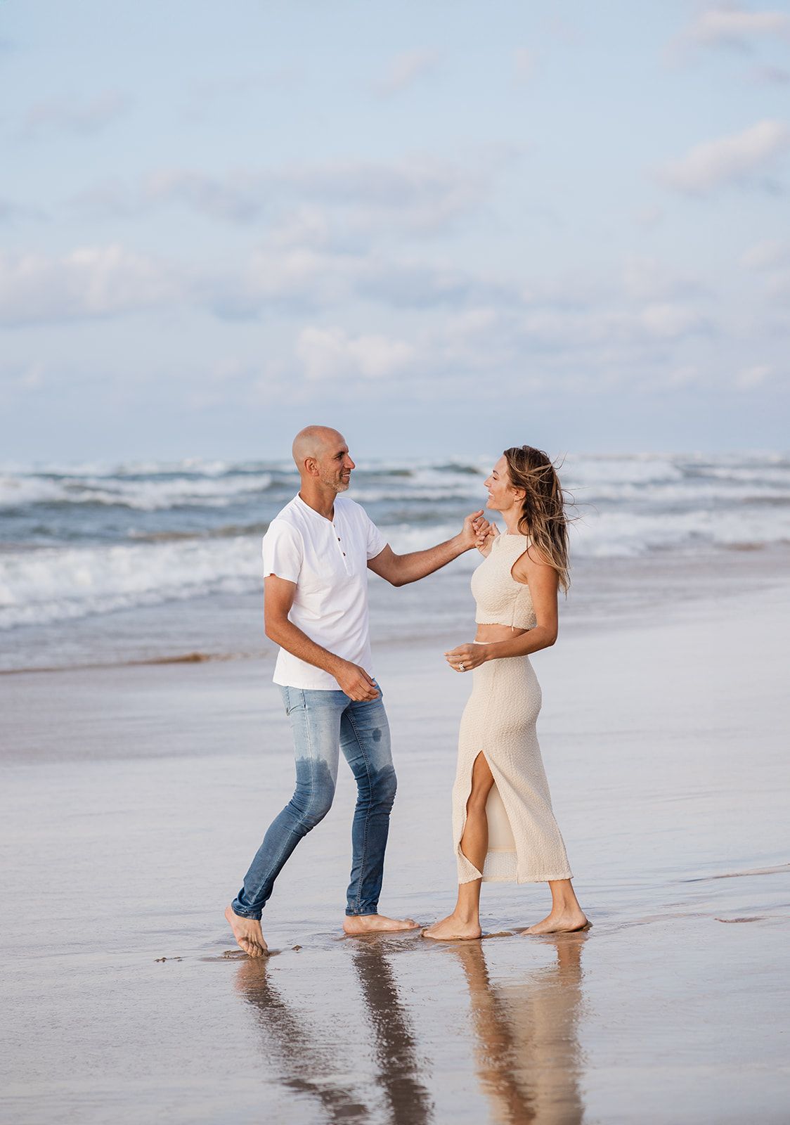 A couple dances barefoot on a sandy beach with ocean waves, man in white shirt and jeans, woman in beige dress.
