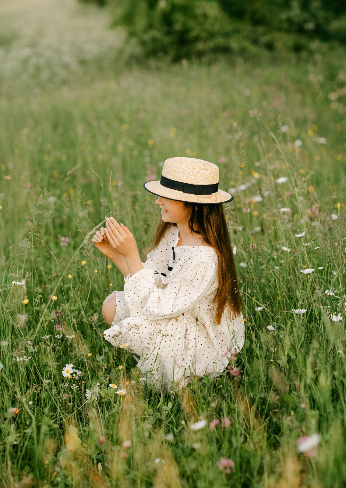 Young girl in straw hat and white floral dress sitting in wildflower meadow holding a flower.