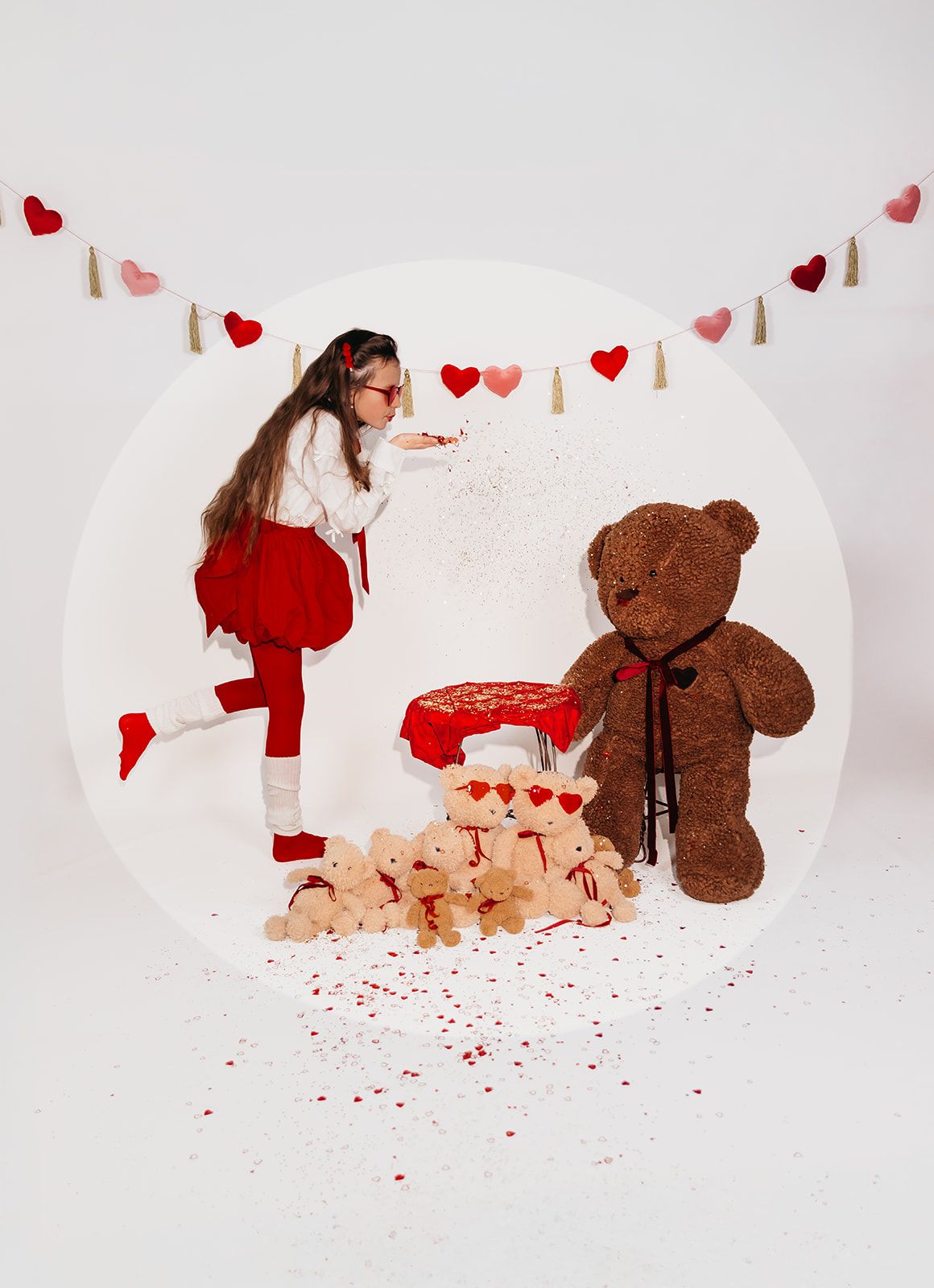 Young girl blowing confetti near teddy bears and heart decorations in Valentine's Day photo setup.