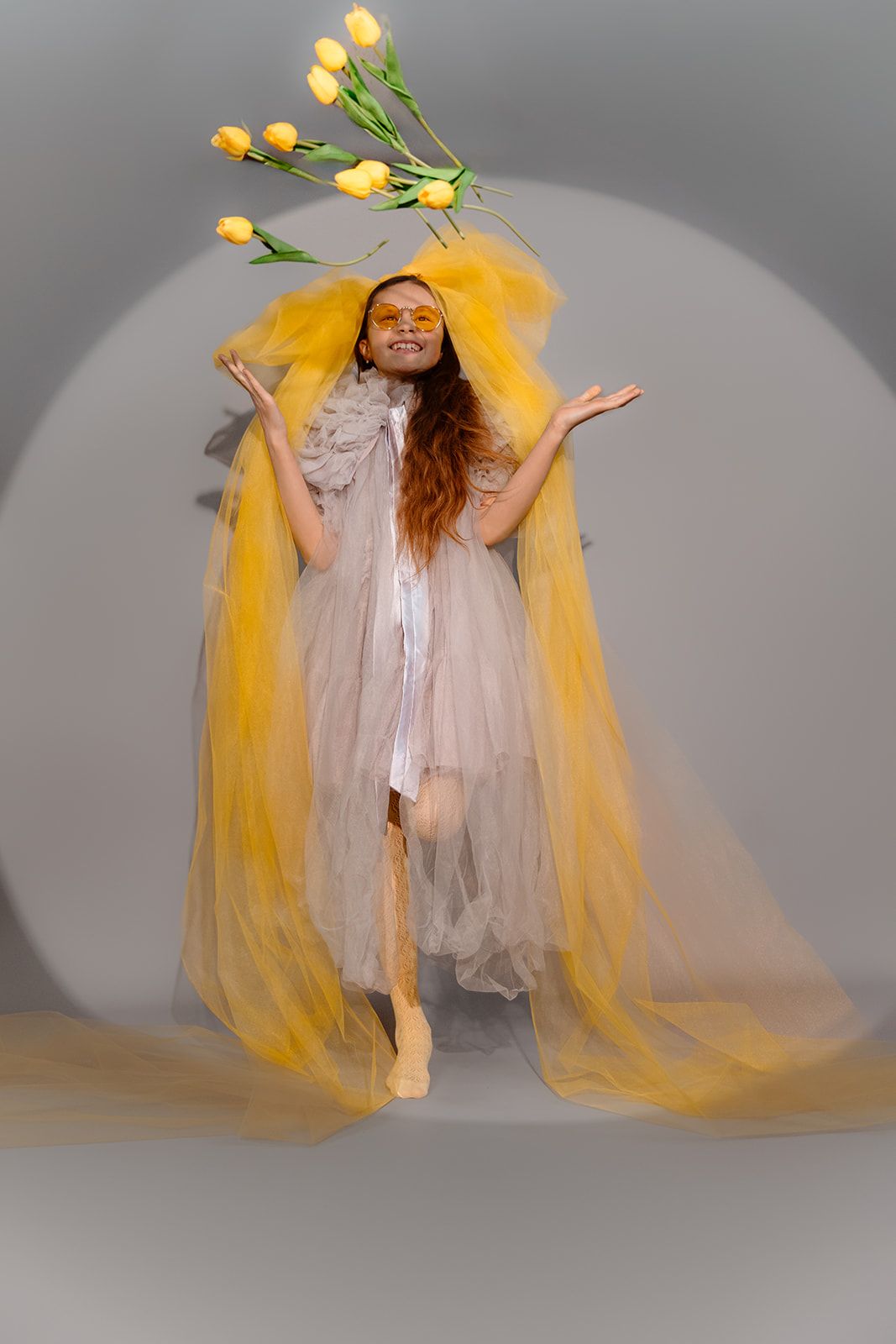 Woman in sheer yellow tulle gown poses with yellow tulips floating above her head on gray background.