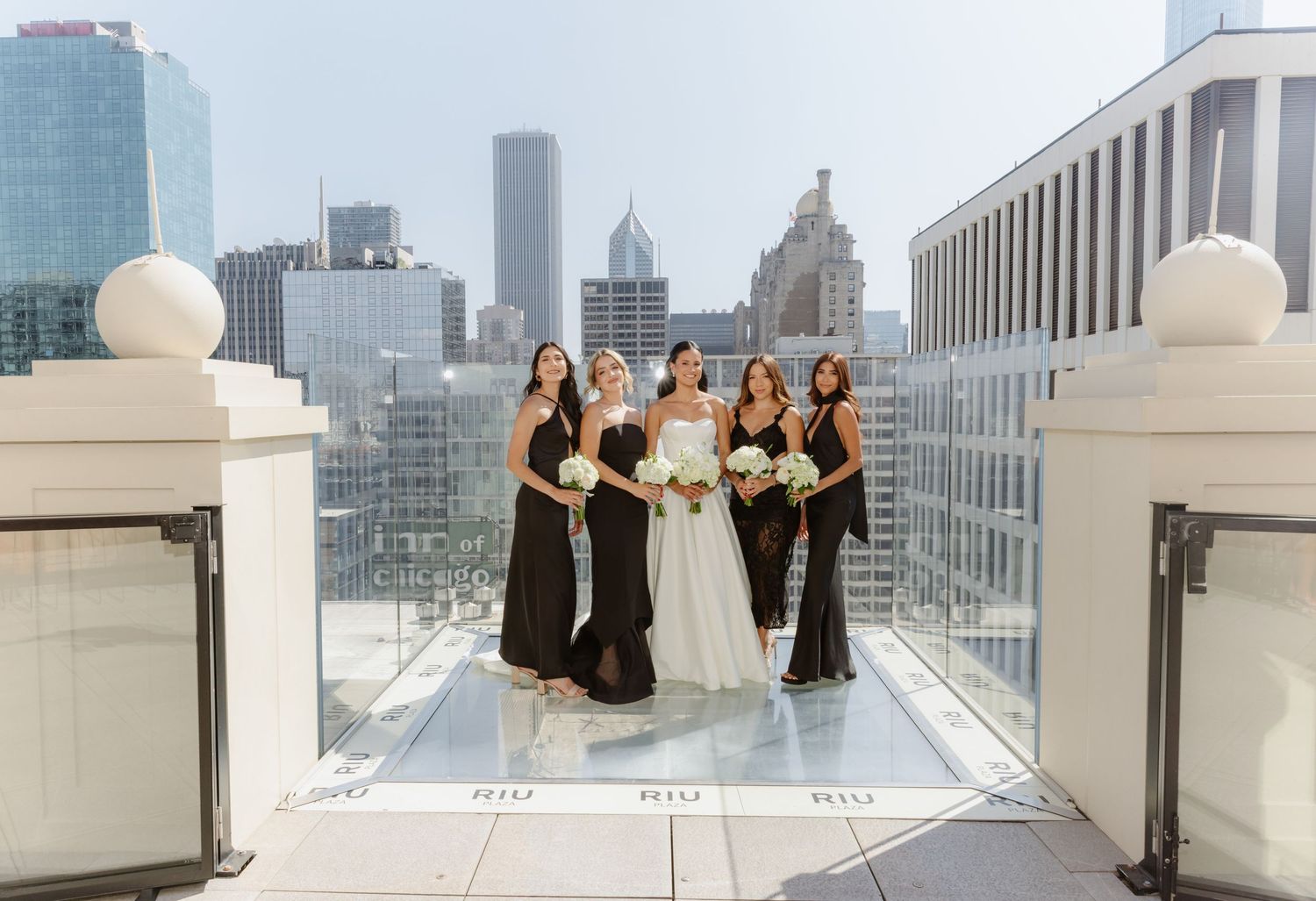 Bride with four bridesmaids in black gowns posing on rooftop terrace with Chicago skyline.