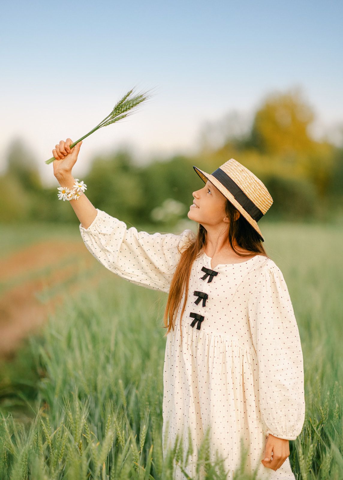 A young girl in a white polka dot dress and straw hat holds wheat stalks in a lush green field at golden hour.