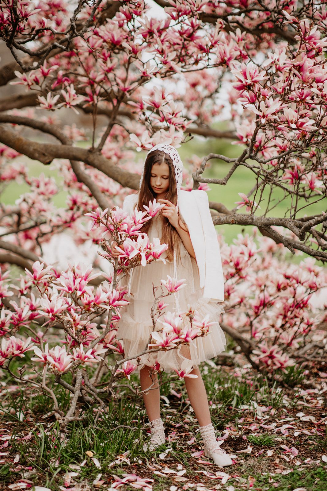 Young girl in white dress holding pink magnolia flowers while standing among blooming magnolia tree branches in spring.