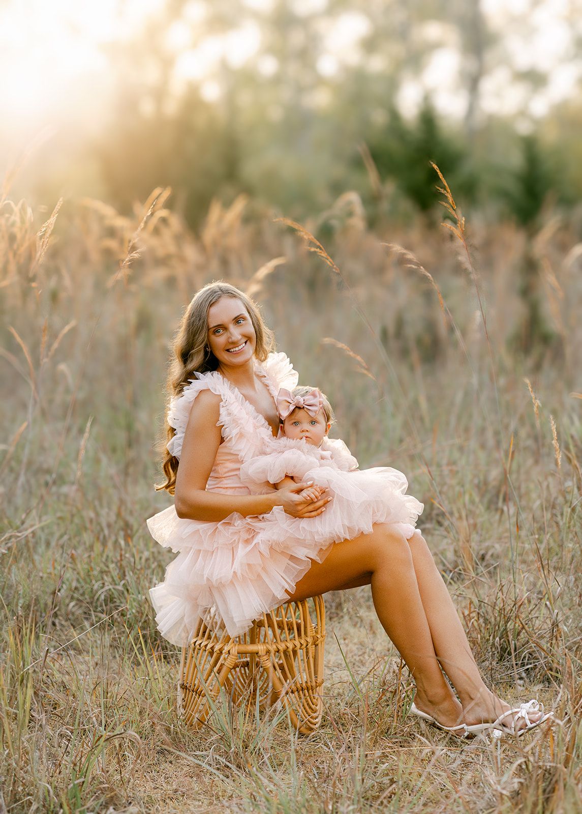 Mother in blush ruffle dress sits on wicker chair holding baby in matching outfit in golden field.