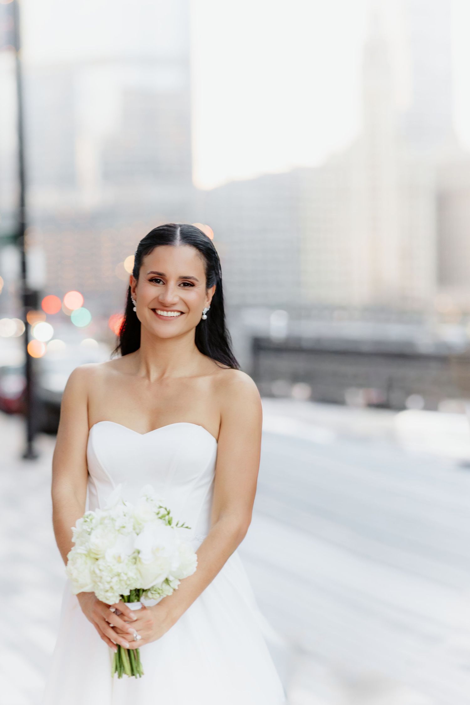 Elegant bride in strapless white ballgown smiles while holding white floral bouquet on a city street at dusk.