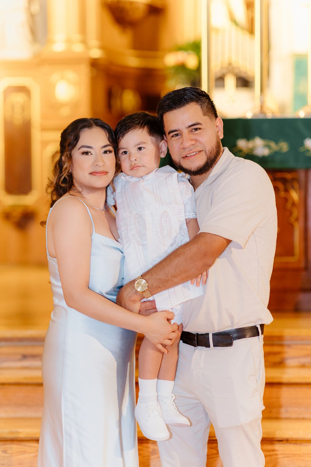 A smiling Hispanic family poses in a church, parents holding their toddler dressed in a white baptism outfit.