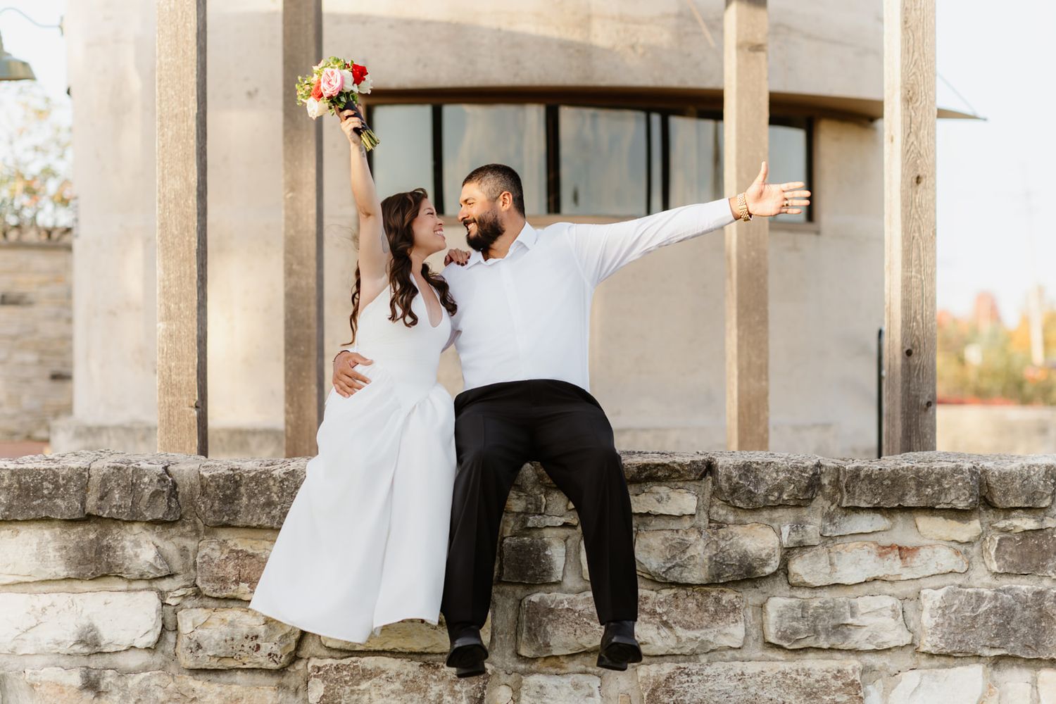 Joyful bride and groom celebrate their wedding day on a stone wall, with the bride raising her colorful bouquet.