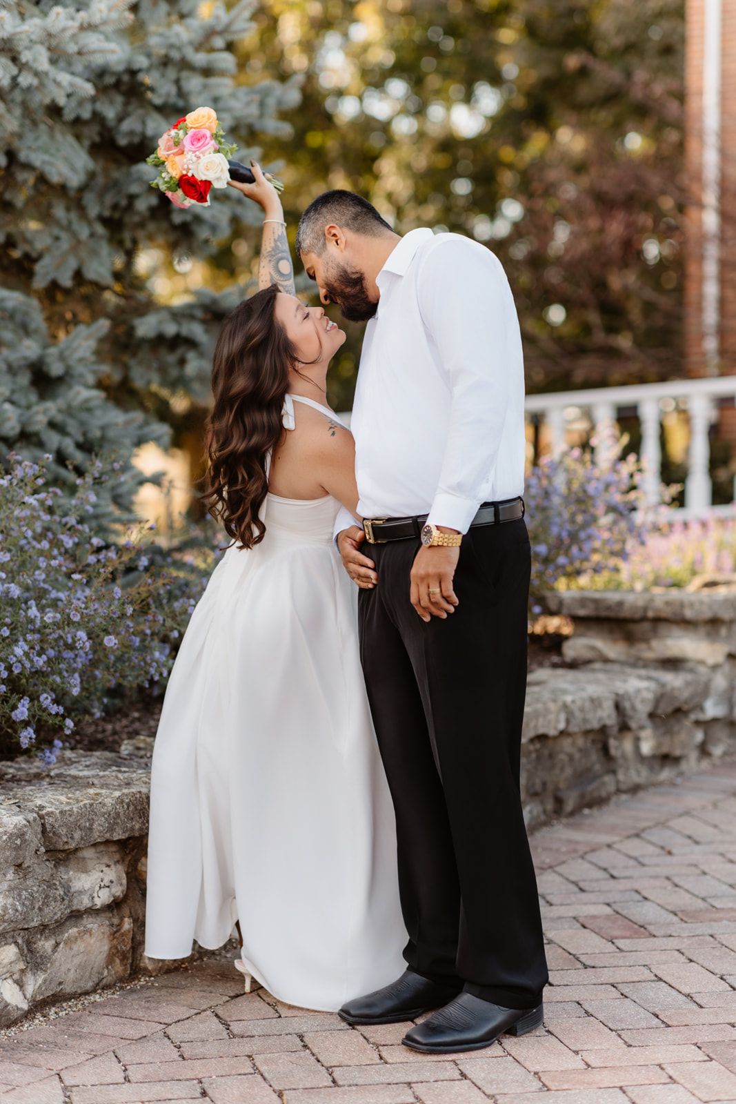 Wedding couple shares romantic kiss outdoors, bride in white dress holds bouquet above while groom wears white shirt.