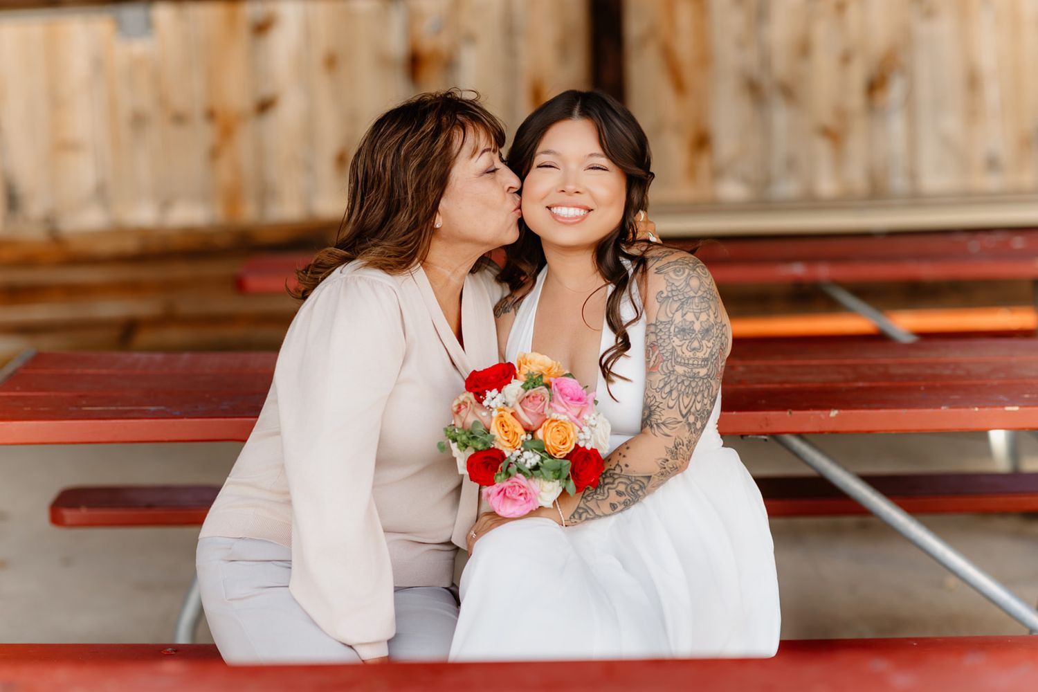 Mother kisses smiling bride on cheek as they sit together holding colorful wedding bouquet.