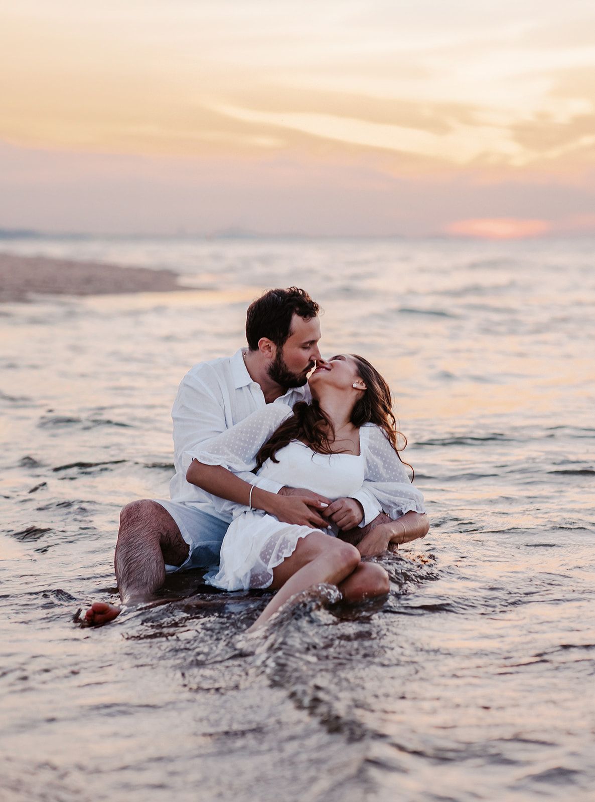 Couple sharing a romantic kiss sitting in ocean waves at sunset, both wearing white outfits on a sandy beach.