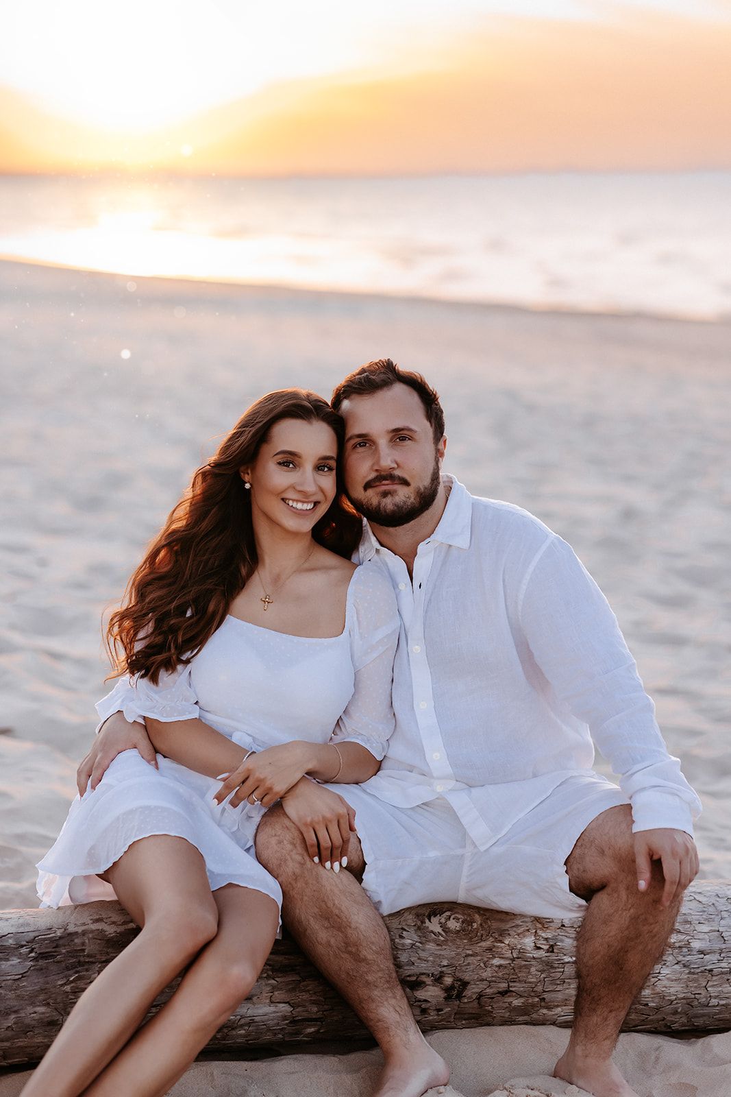 Couple in white outfits sitting on driftwood at a sandy beach during a golden sunset.