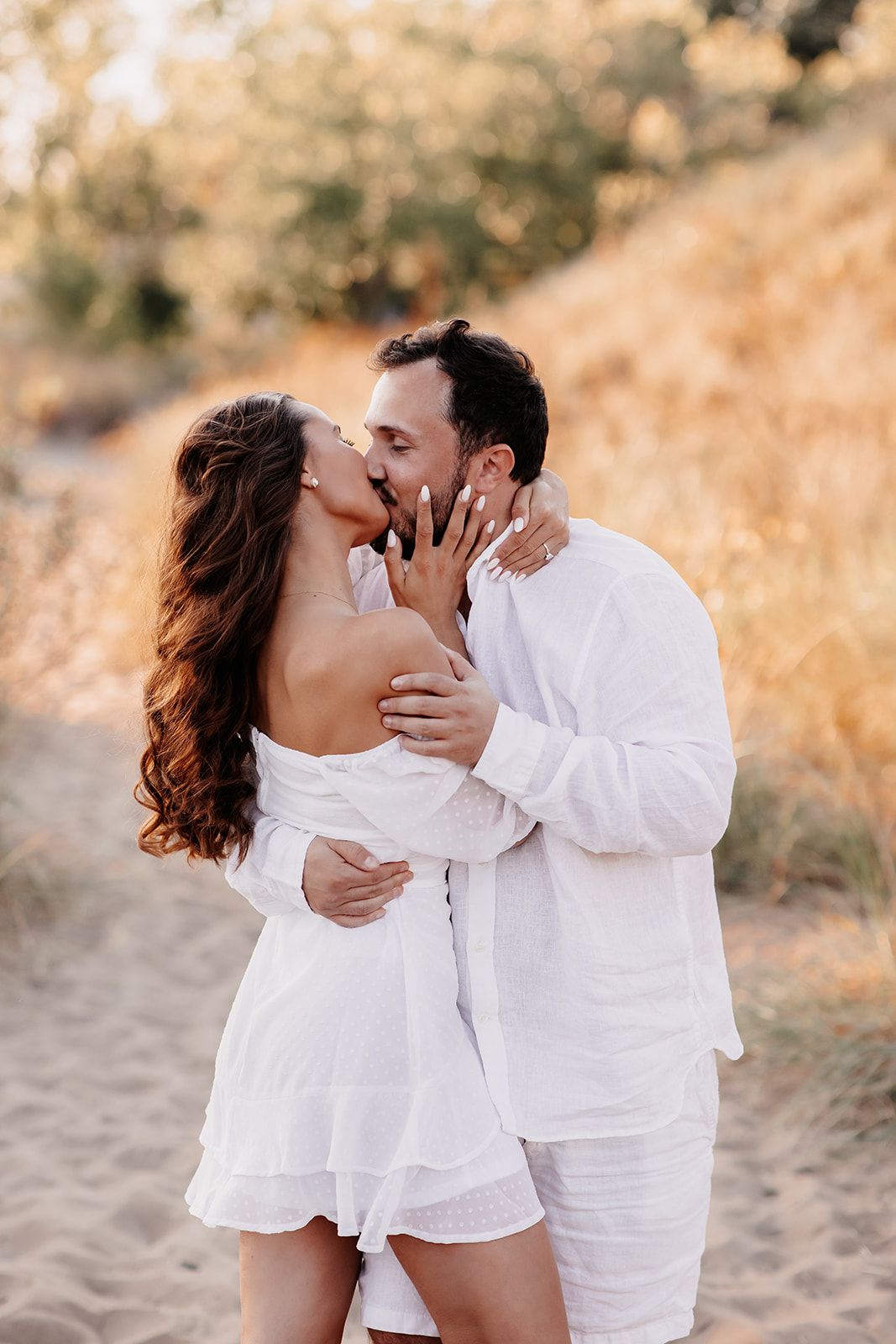 Couple in white outfits sharing a romantic kiss on a sandy beach path surrounded by golden grasses.