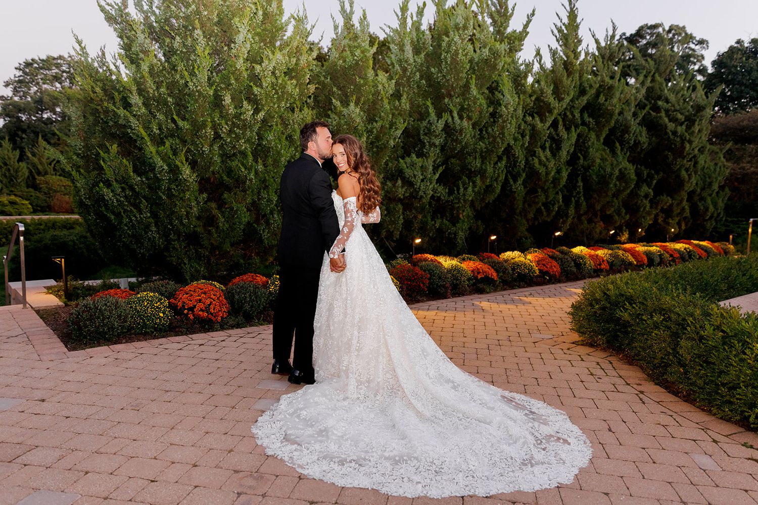Bride and groom share a romantic kiss on a brick path surrounded by tall evergreens and colorful fall flowers.