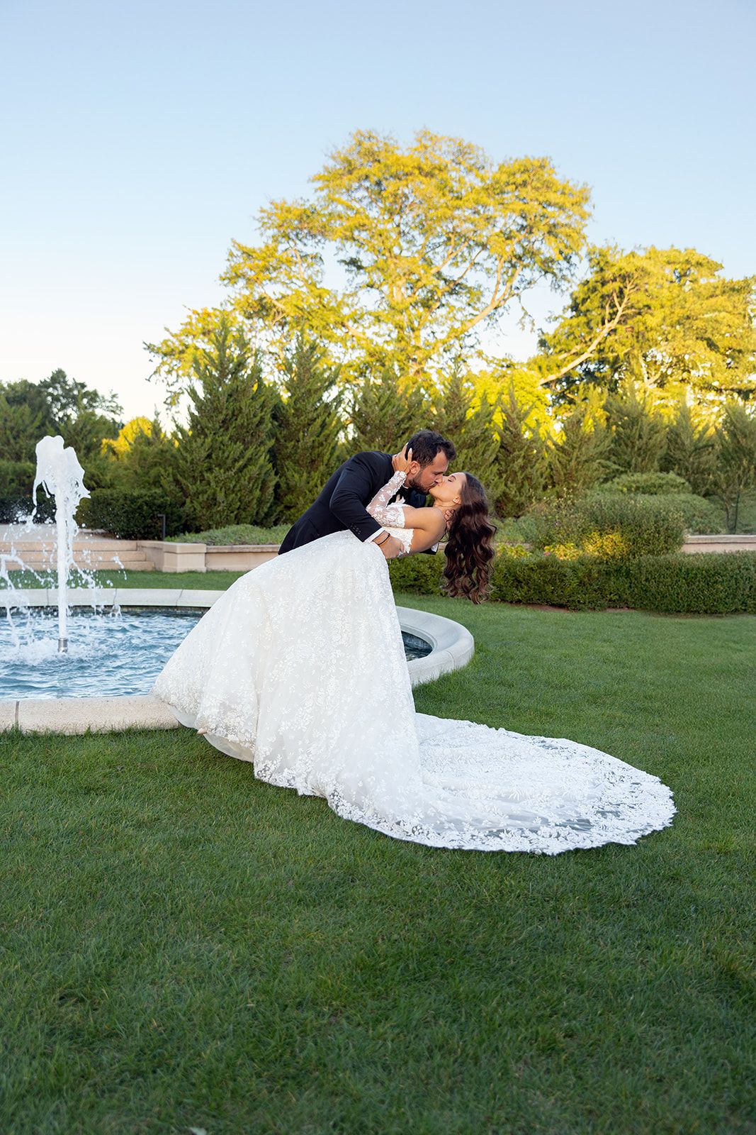 Bride and groom share a romantic dip kiss in front of a garden fountain, bride in elegant lace gown.