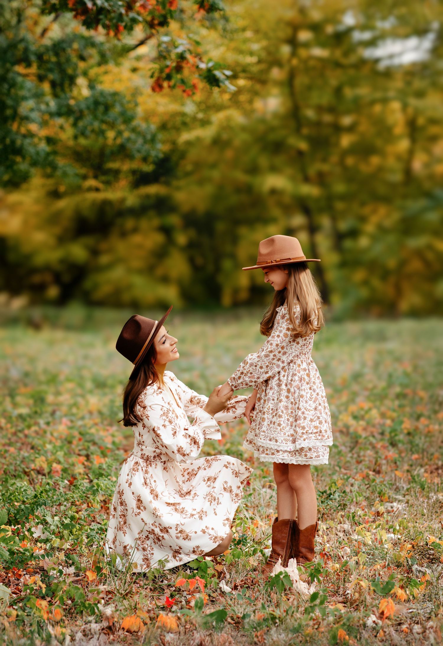 A mother and daughter in matching floral dresses and brown hats hold hands in an autumn field.