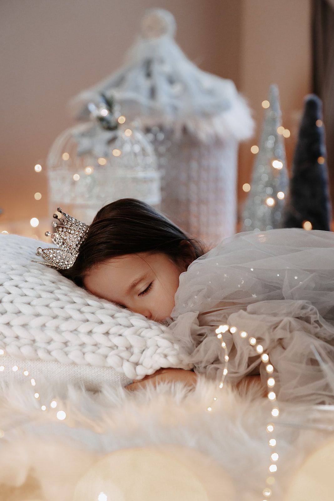 Young girl wearing a silver crown sleeping peacefully on white knit pillow surrounded by glowing Christmas lights and decorations.