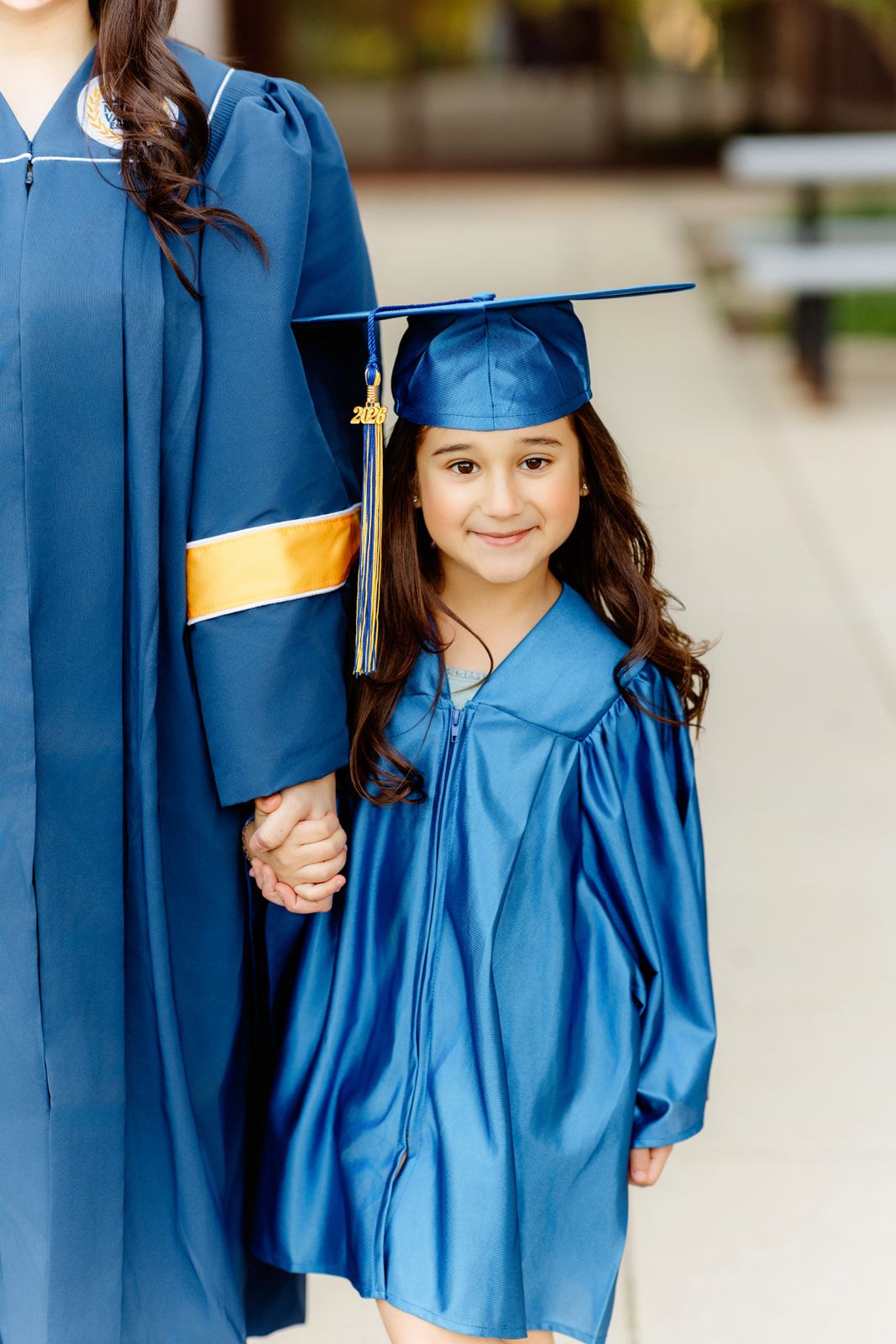 Young girl in shiny blue graduation cap and gown holding hands with older graduate on walkway.