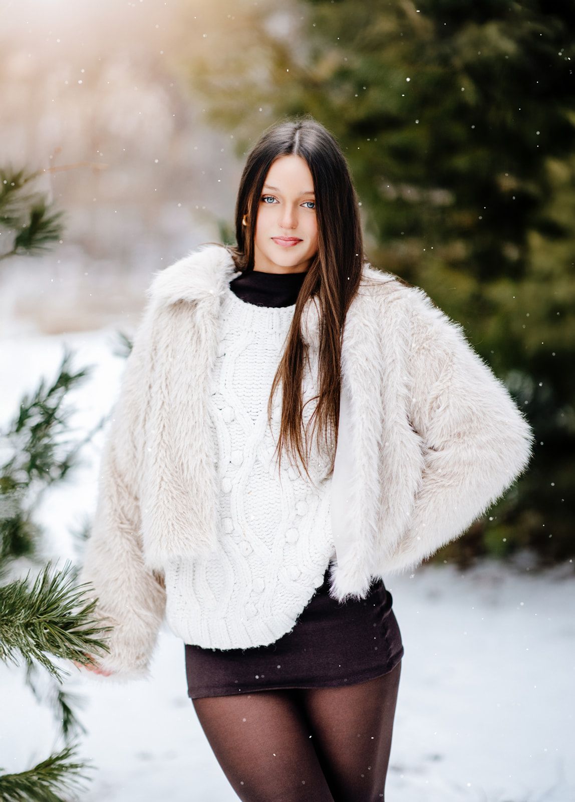 Smiling young brunette woman in white faux fur jacket and black mini skirt posing near pine trees in falling snow.
