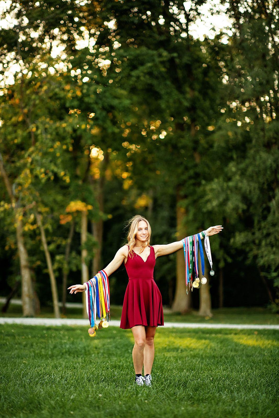 Blonde girl in red dress displaying colorful medals with arms outstretched in a green park.