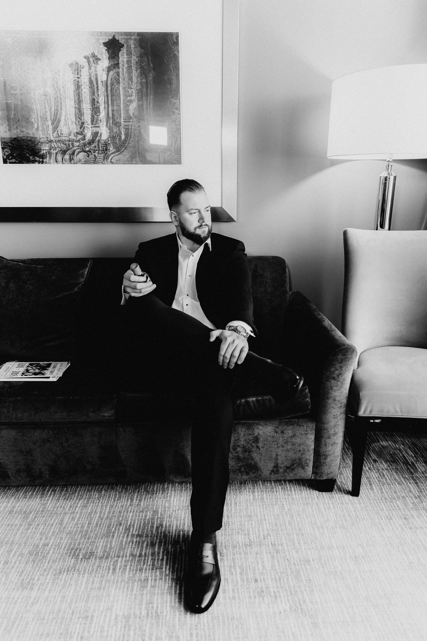 Groom in black suit holding drink while seated on sofa, black and white hotel room portrait.