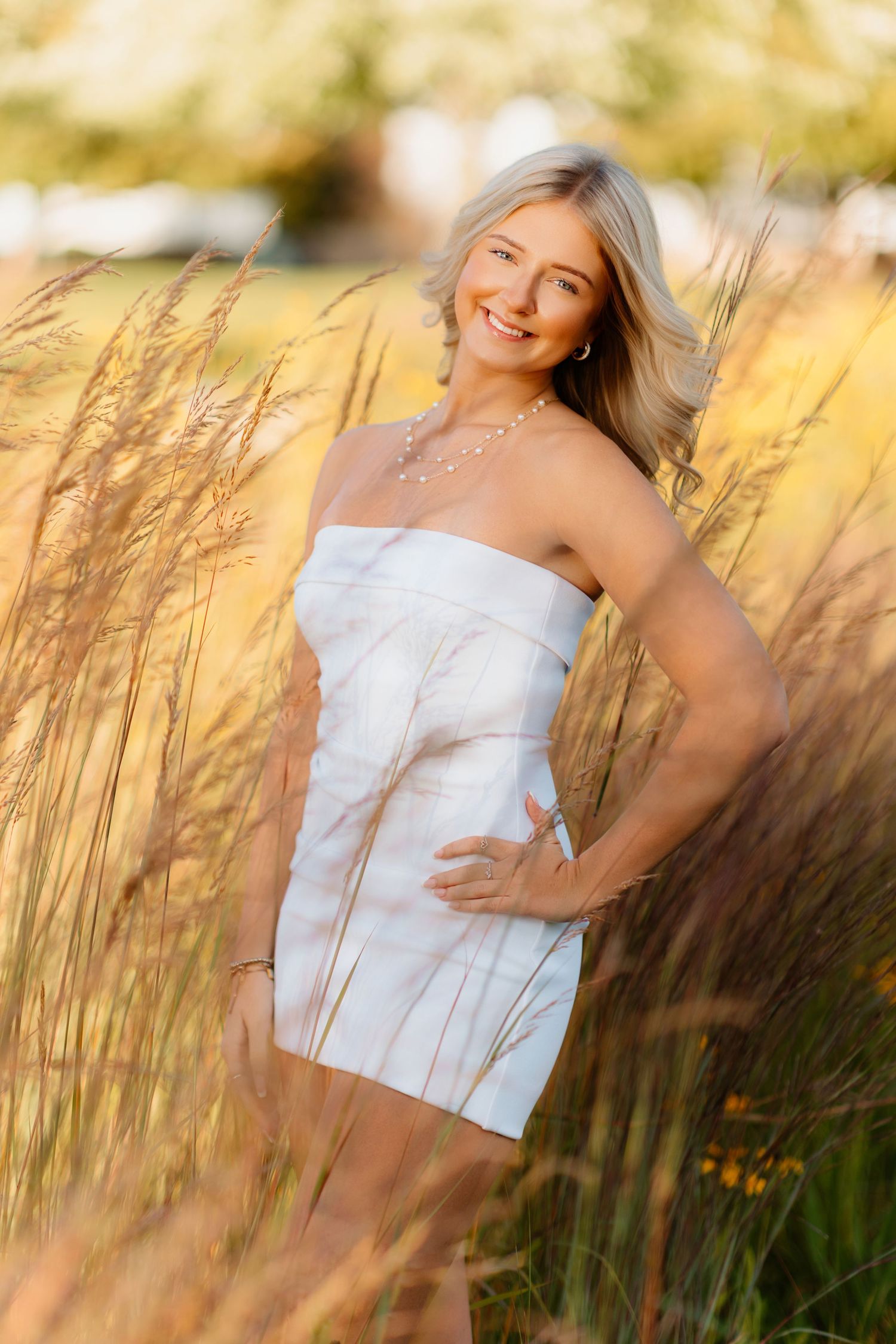 Smiling blonde woman in white strapless dress standing among tall golden grass outdoors.