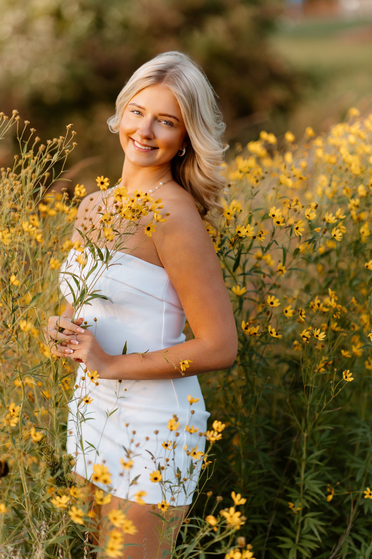 Blonde woman in white strapless dress smiling among golden yellow wildflowers at sunset.