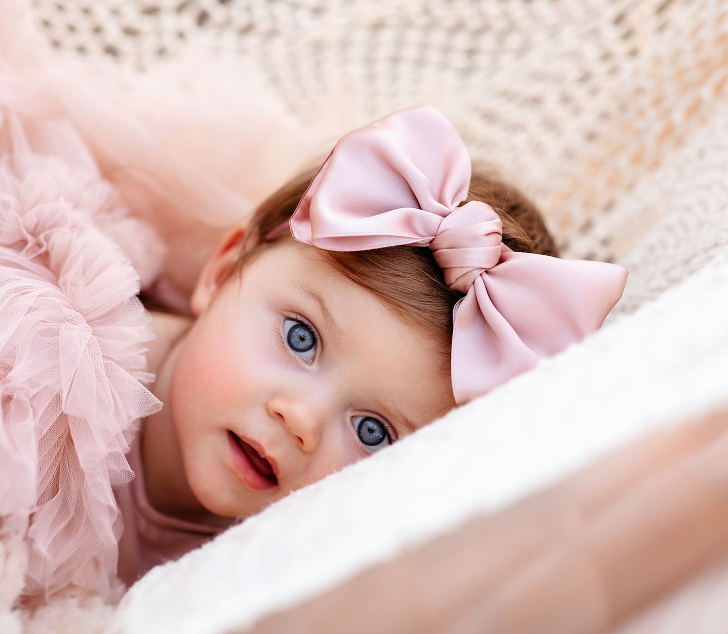 Close-up of adorable blue-eyed baby girl wearing pink bow headband lying in wicker basket.