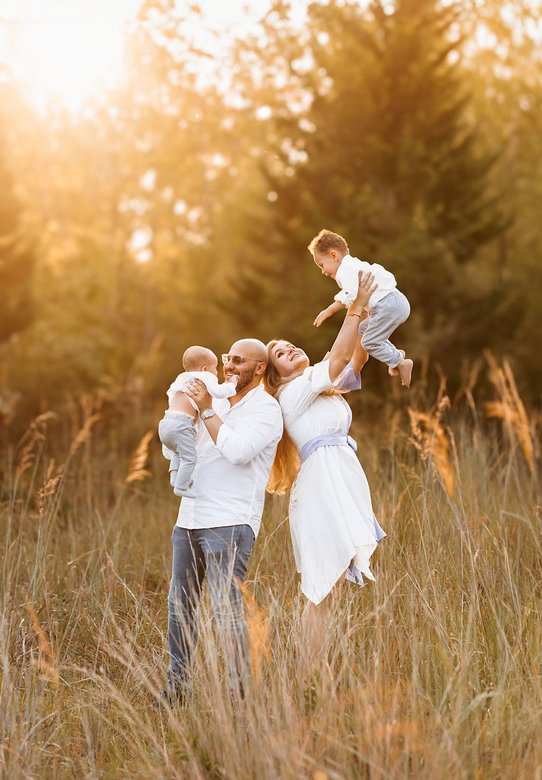 A joyful family plays in a sunlit meadow, with the mother lifting a toddler while father holds a baby.