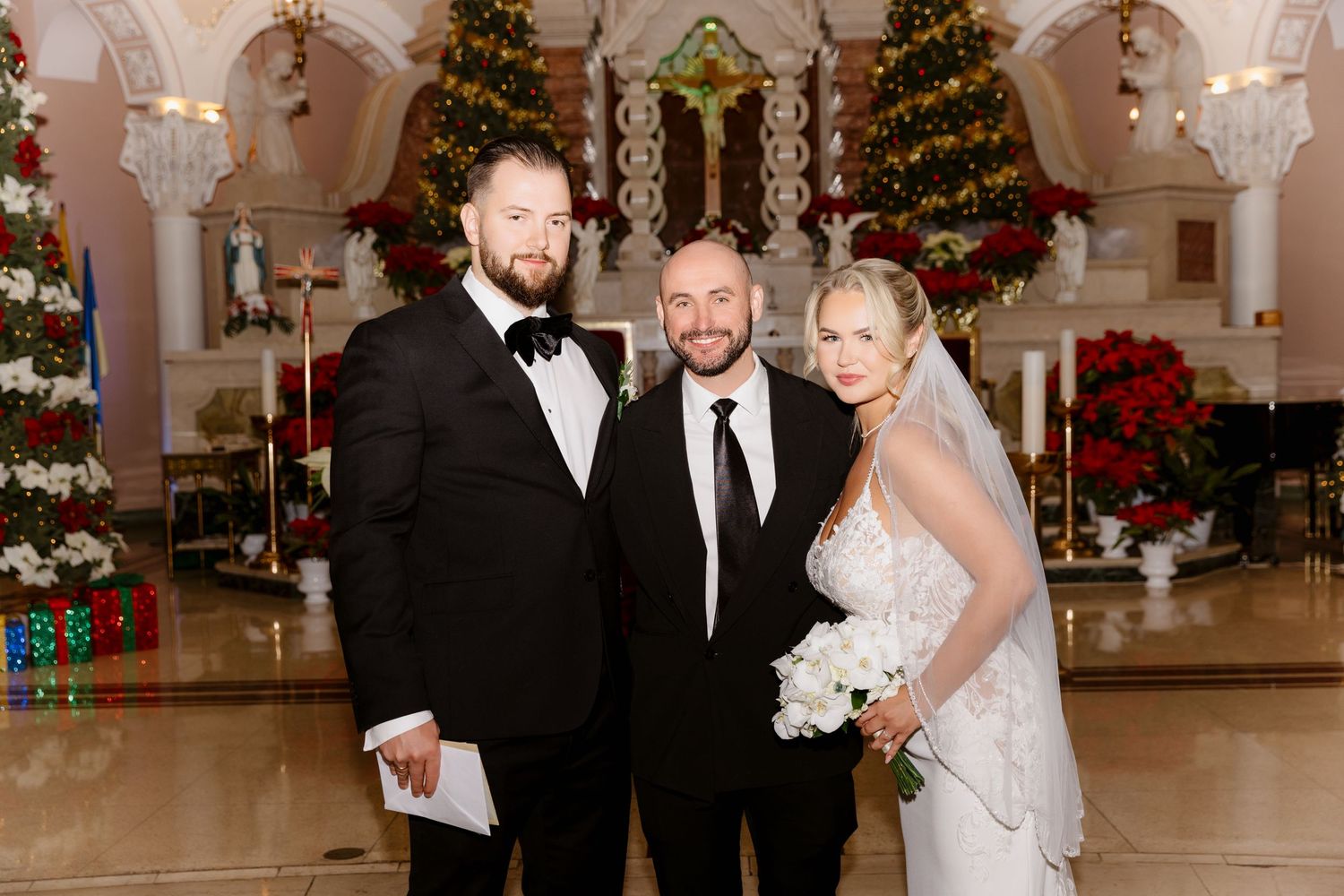 Bride groom and male guest smiling together in Christmas-decorated church with holiday decor behind.