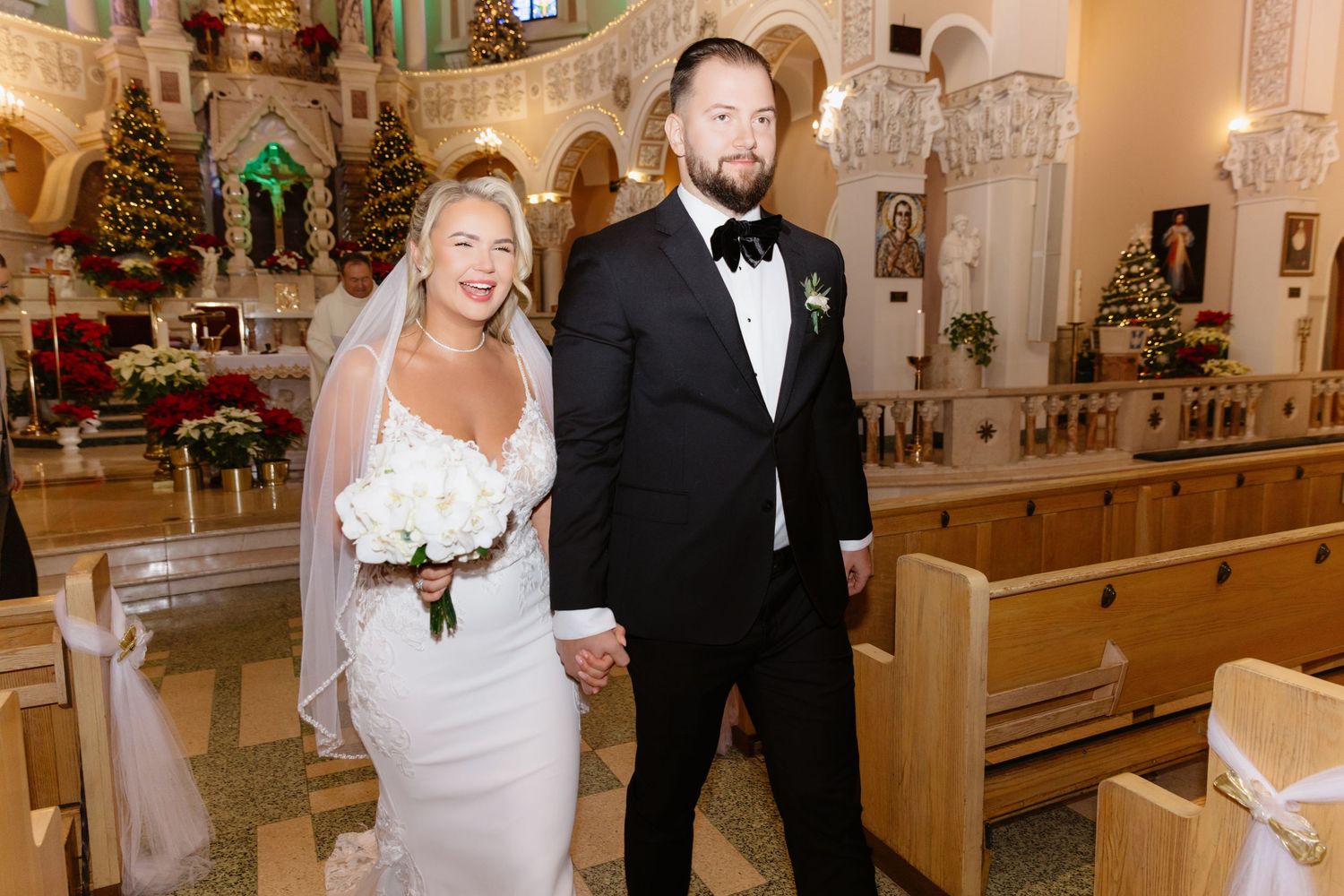 Bride holding white bouquet and groom in black tuxedo pose together in Christmas-decorated church aisle.