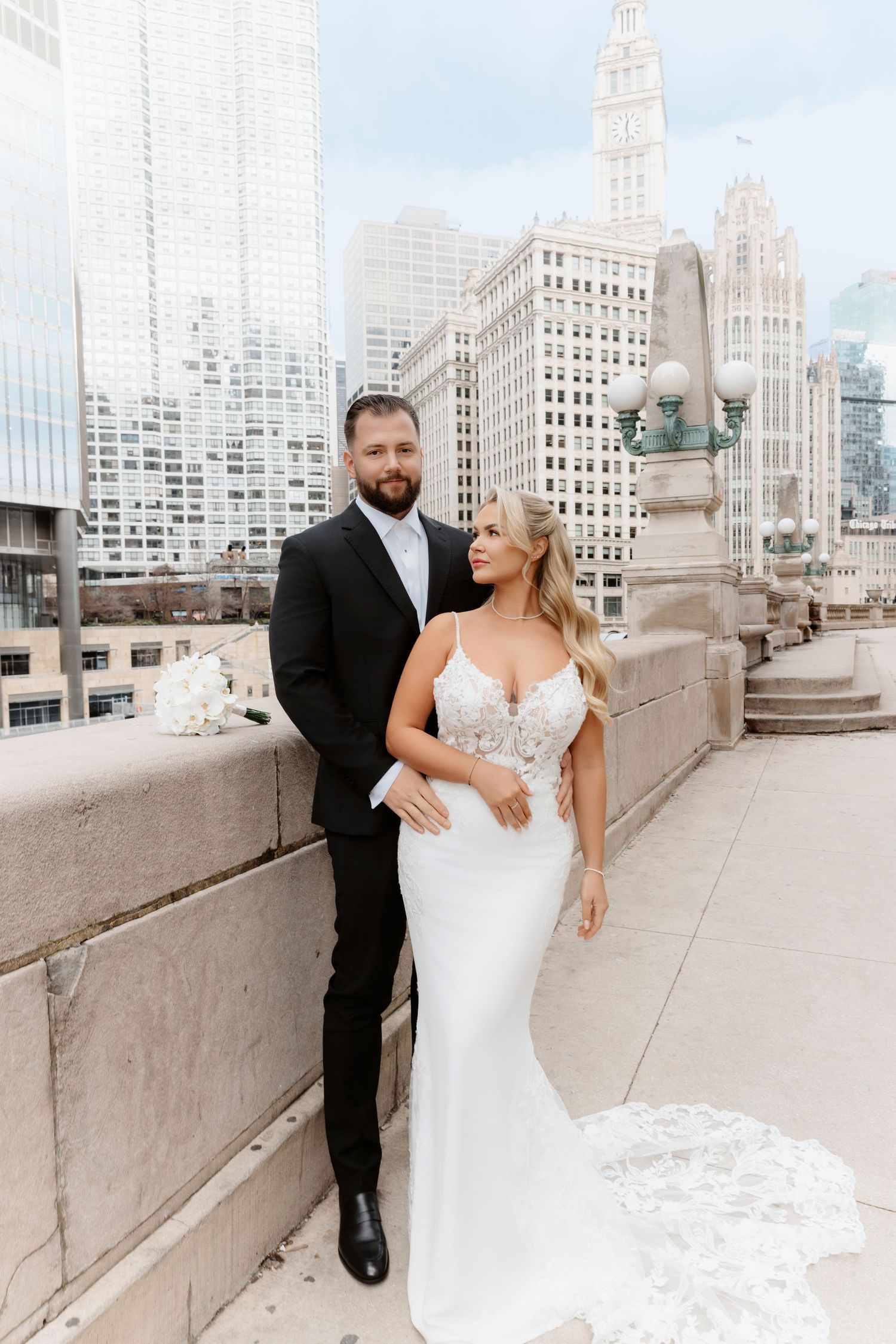 Bride in spaghetti strap lace gown poses with groom on Chicago bridge with iconic Tribune Tower behind.
