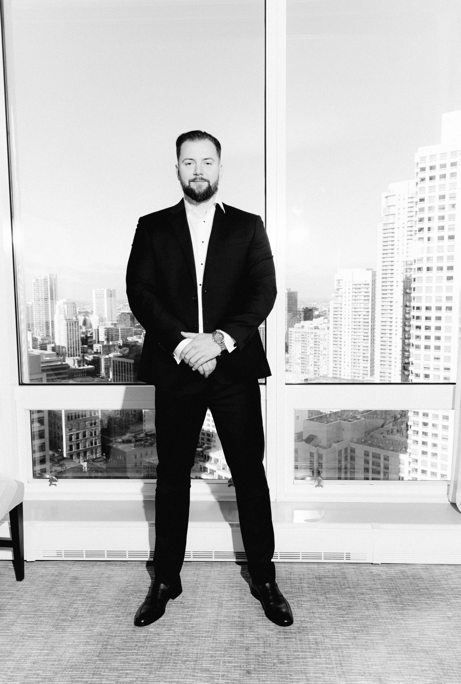 Groom in black suit stands confidently before city skyline window in black and white wedding portrait.