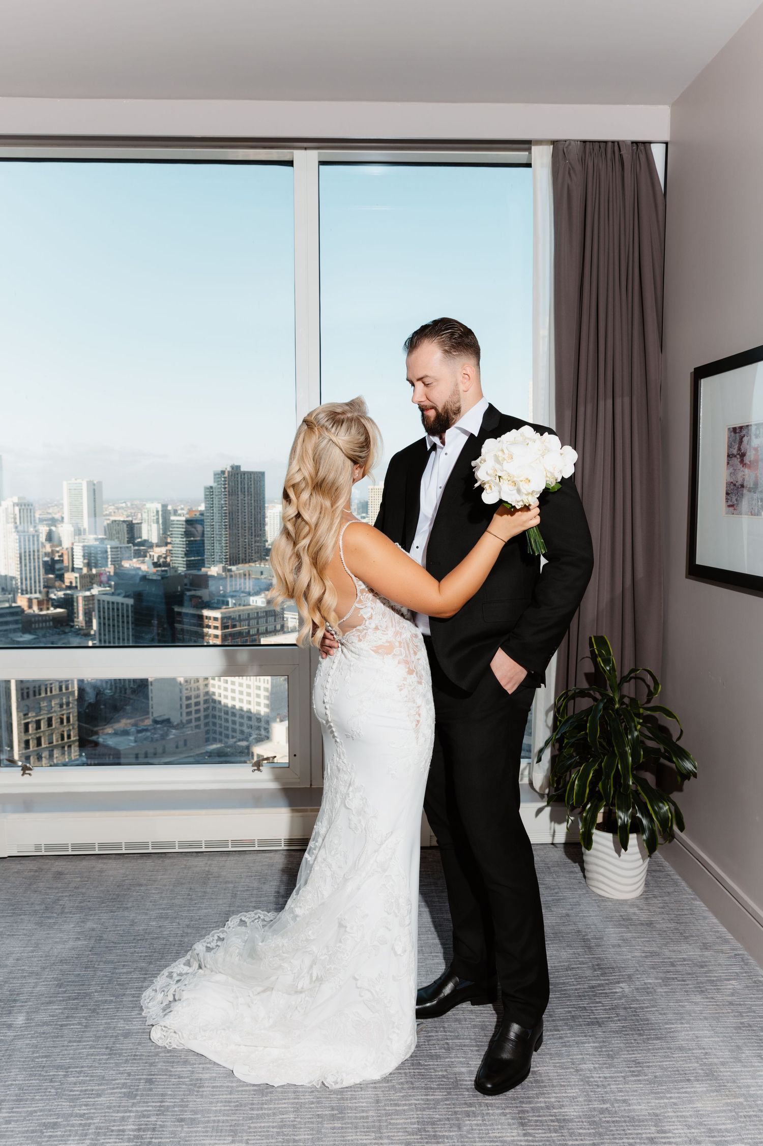 Bride in lace gown faces groom holding white bouquet during intimate first look in hotel room.
