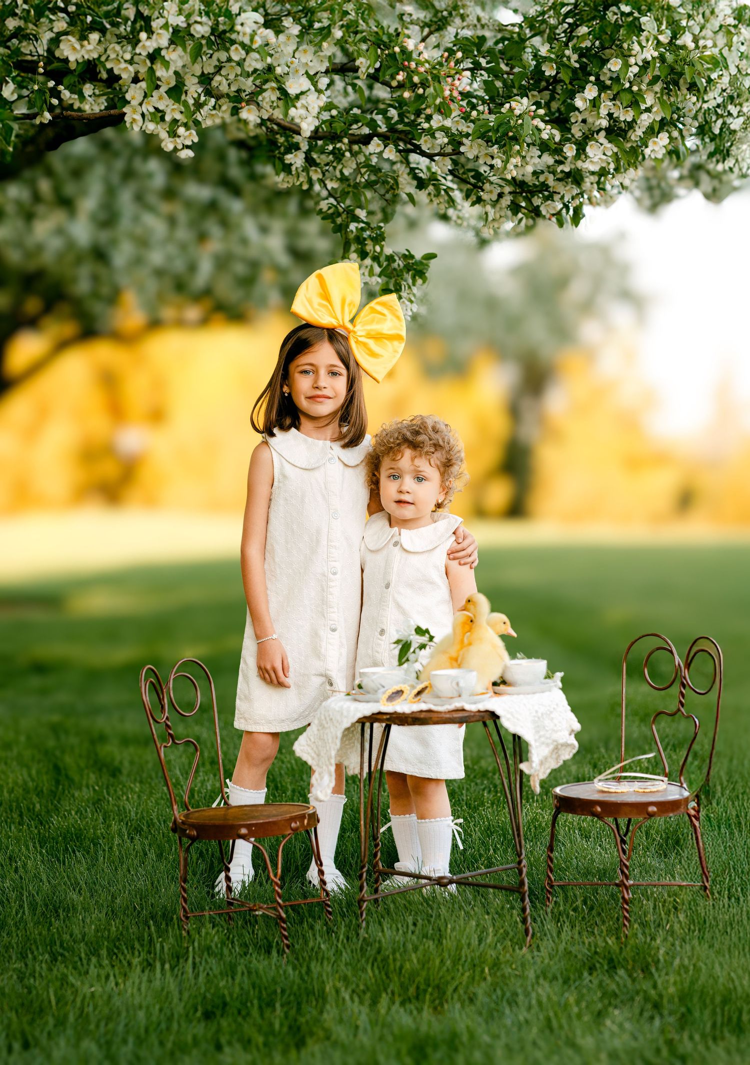 Two young girls in white dresses pose outdoors at a miniature tea party under blooming white trees on lush green grass.