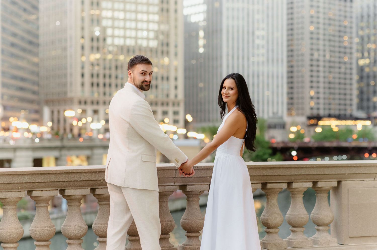 Elegant couple holding hands on bridge overlooking Chicago River with glowing city lights.