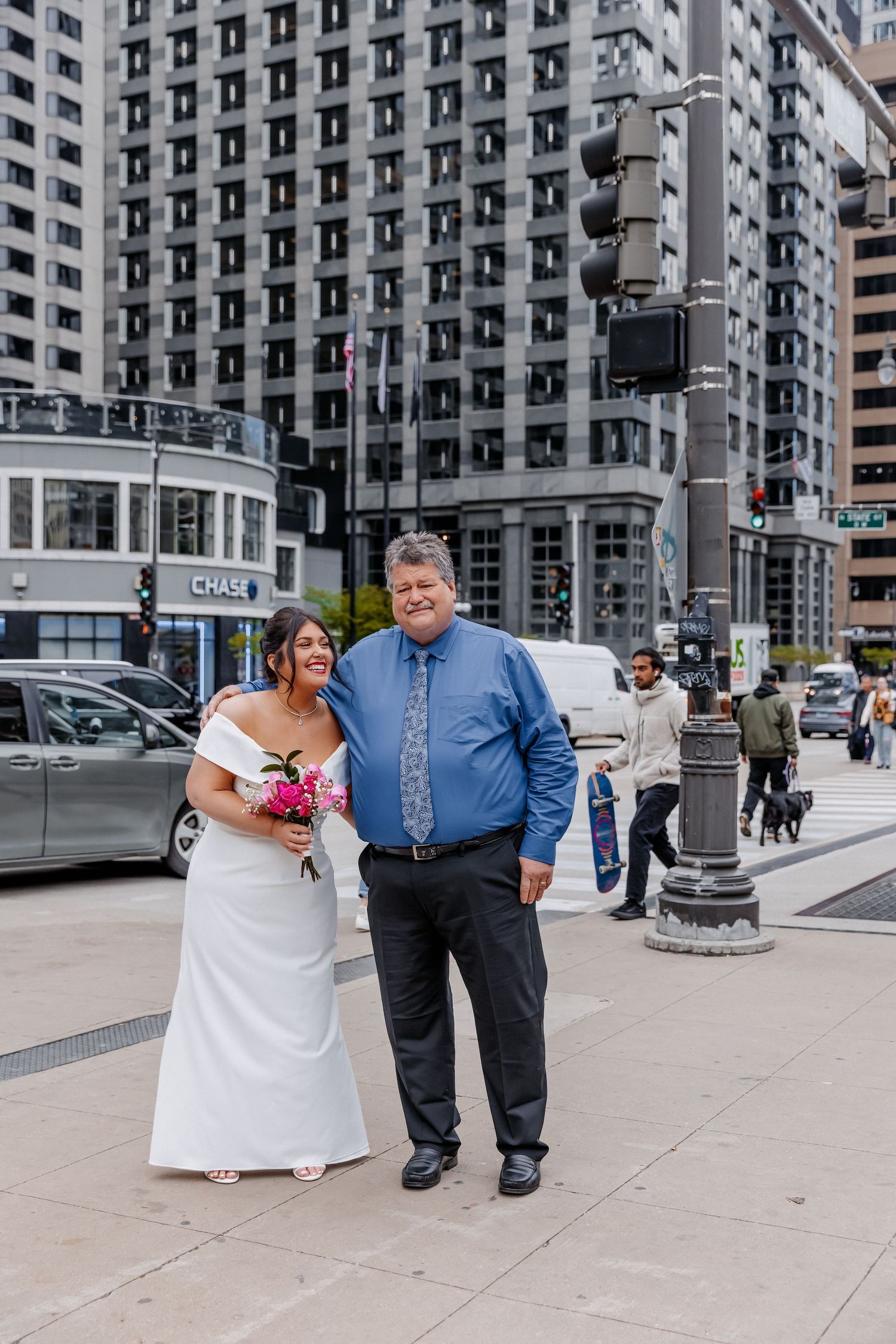 Bride in white dress and groom in blue shirt smile together on a busy city street corner.