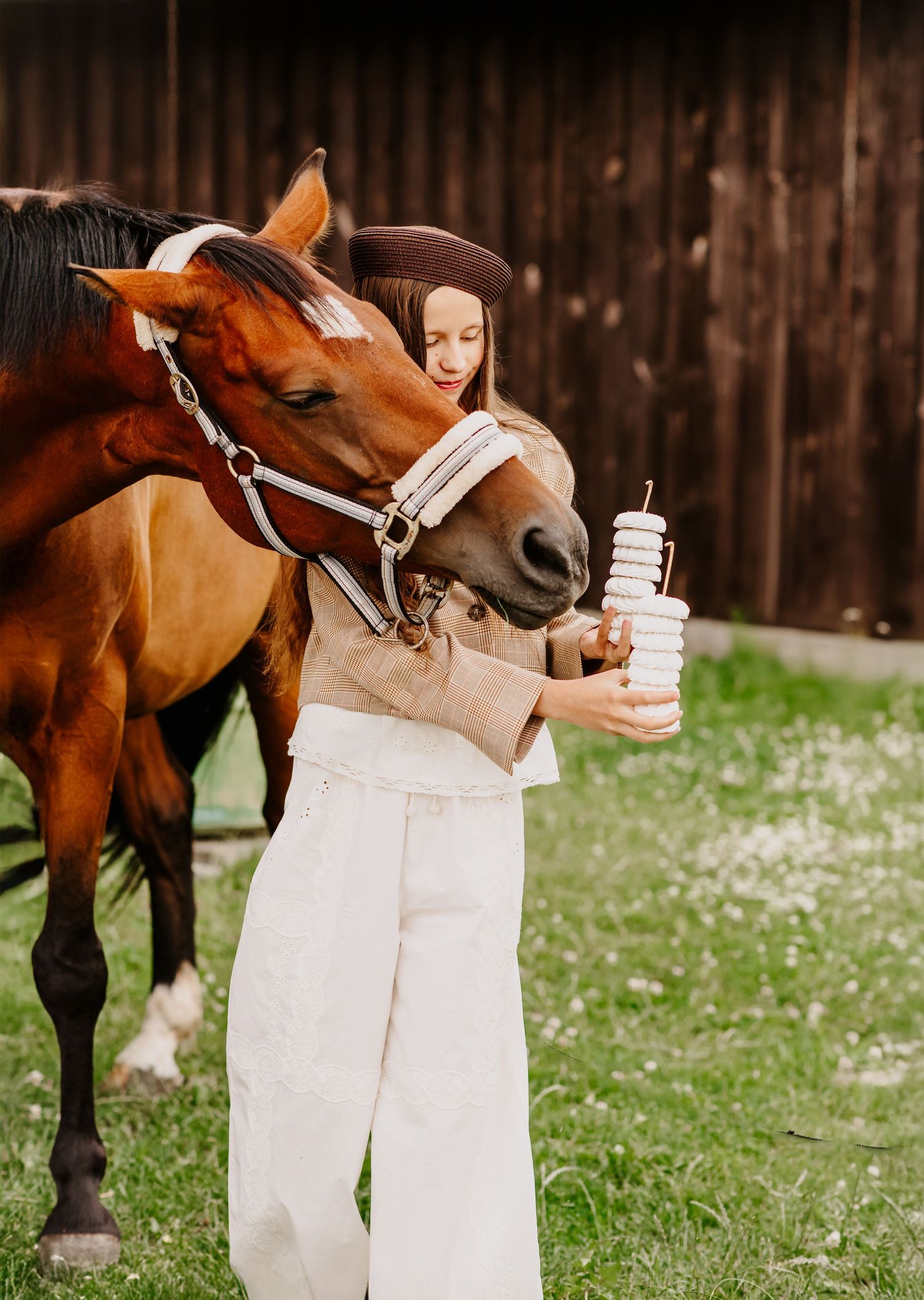 Young woman in beret holds stacked donuts while a chestnut horse sniffs them near a barn.