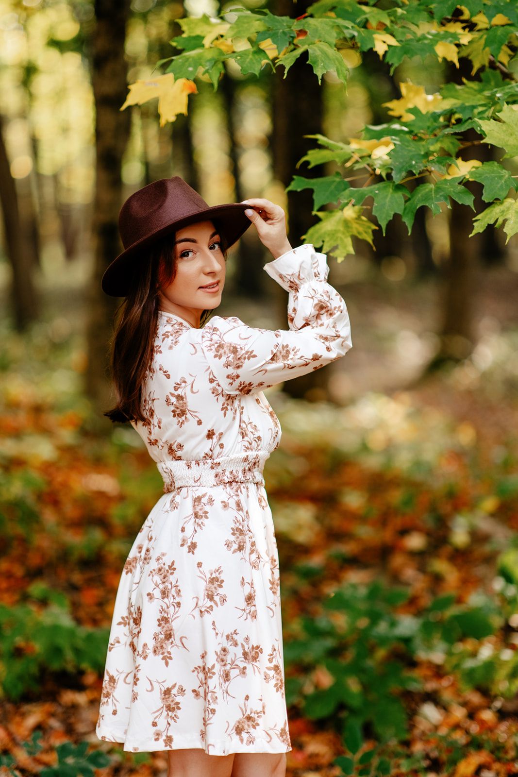 Woman in floral dress and brown hat posing in autumn forest with colorful fallen leaves.