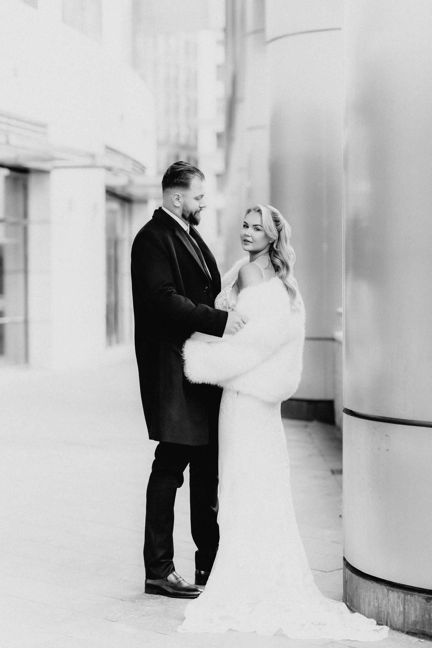 Black and white wedding portrait of couple embracing near architectural column in urban setting.
