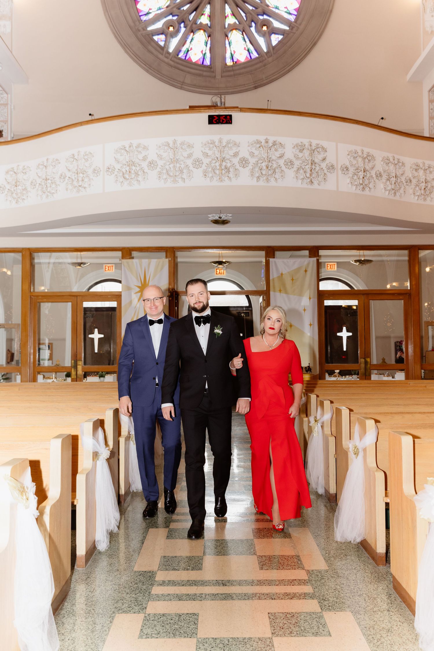 Groom walks down church aisle escorted by parents in blue suit and red dress during wedding processional.