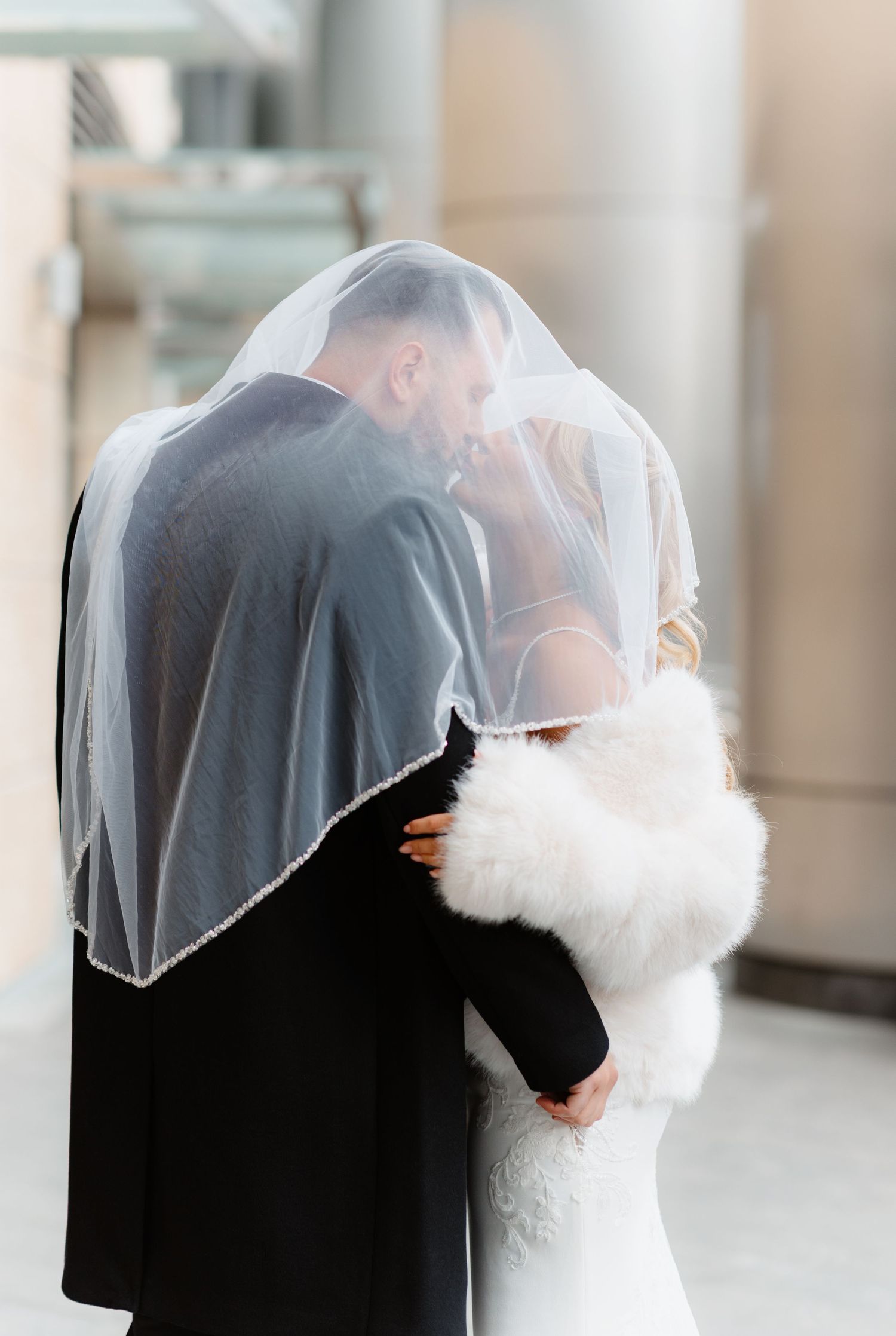 Bride and groom share romantic kiss beneath flowing wedding veil outdoors.