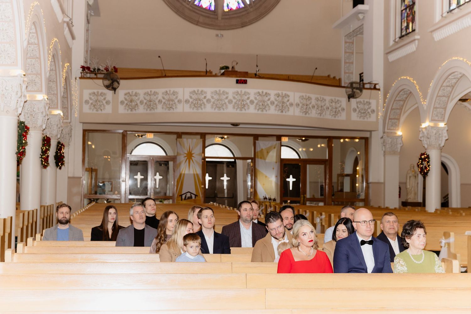 Wedding guests seated in ornate white church pews during formal ceremony with balcony view.