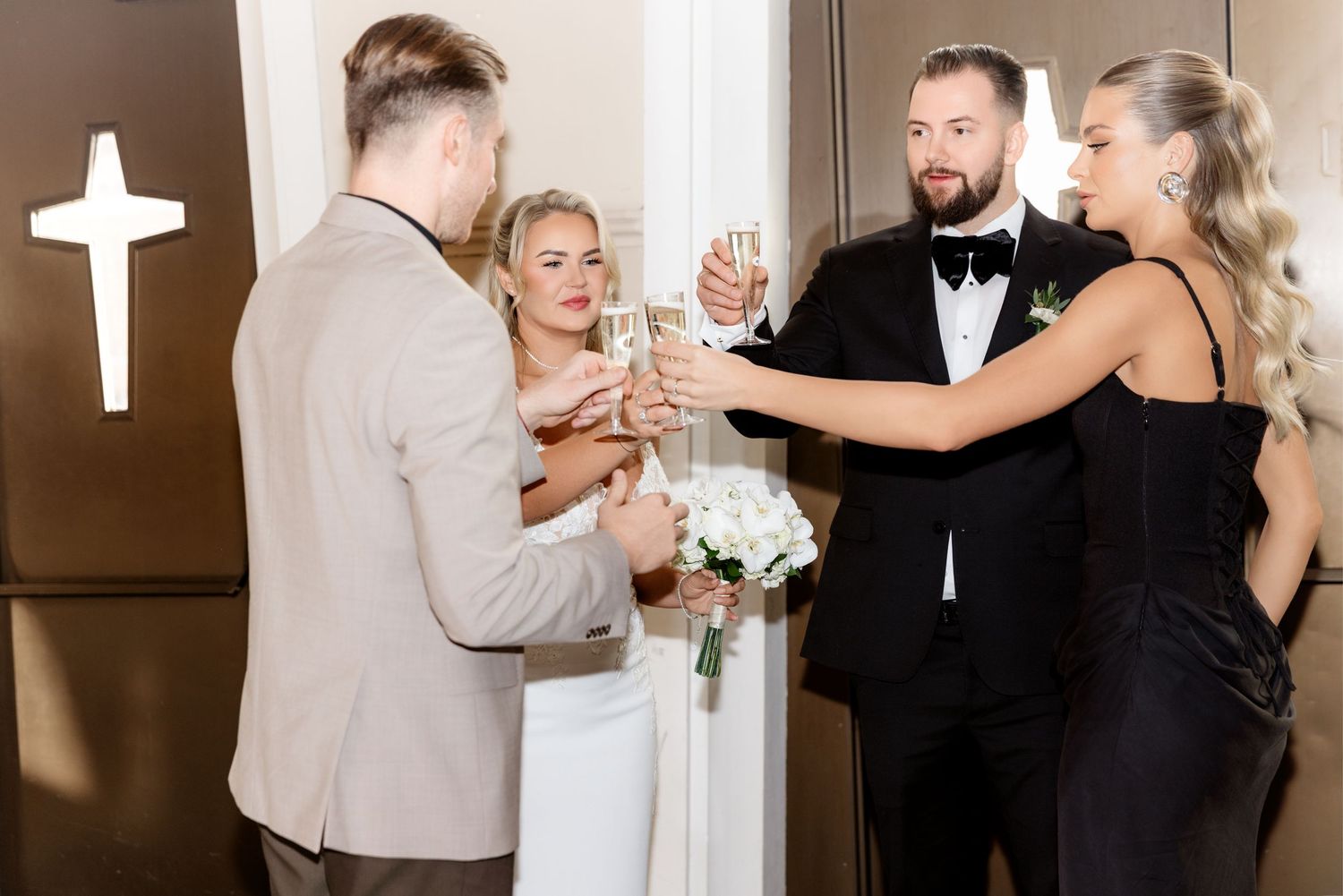 Wedding party toasting with champagne glasses near a cross-adorned door, bride in white gown and guests in formal attire.