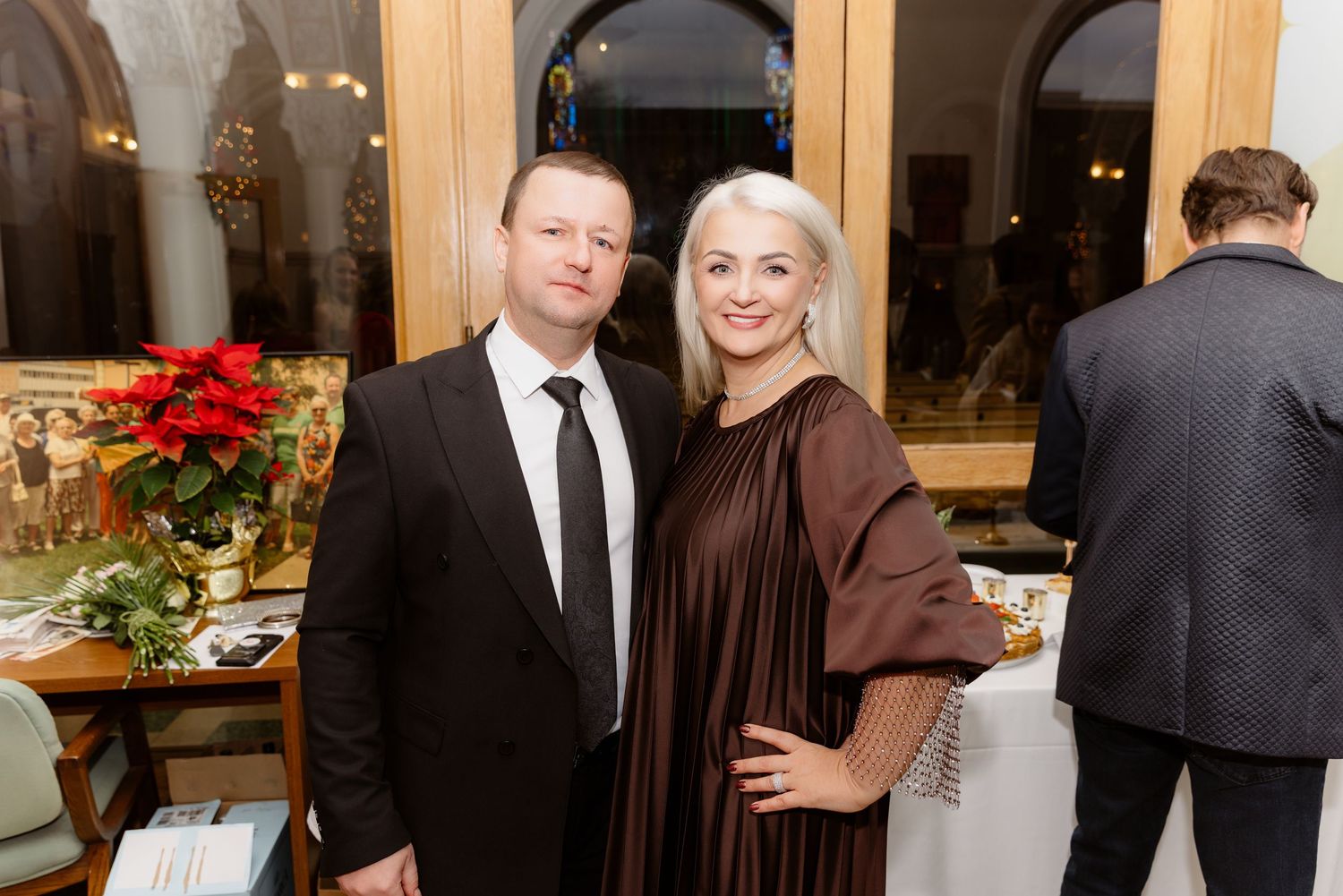Elegant couple posing at formal event, man in black suit and woman in brown dress.