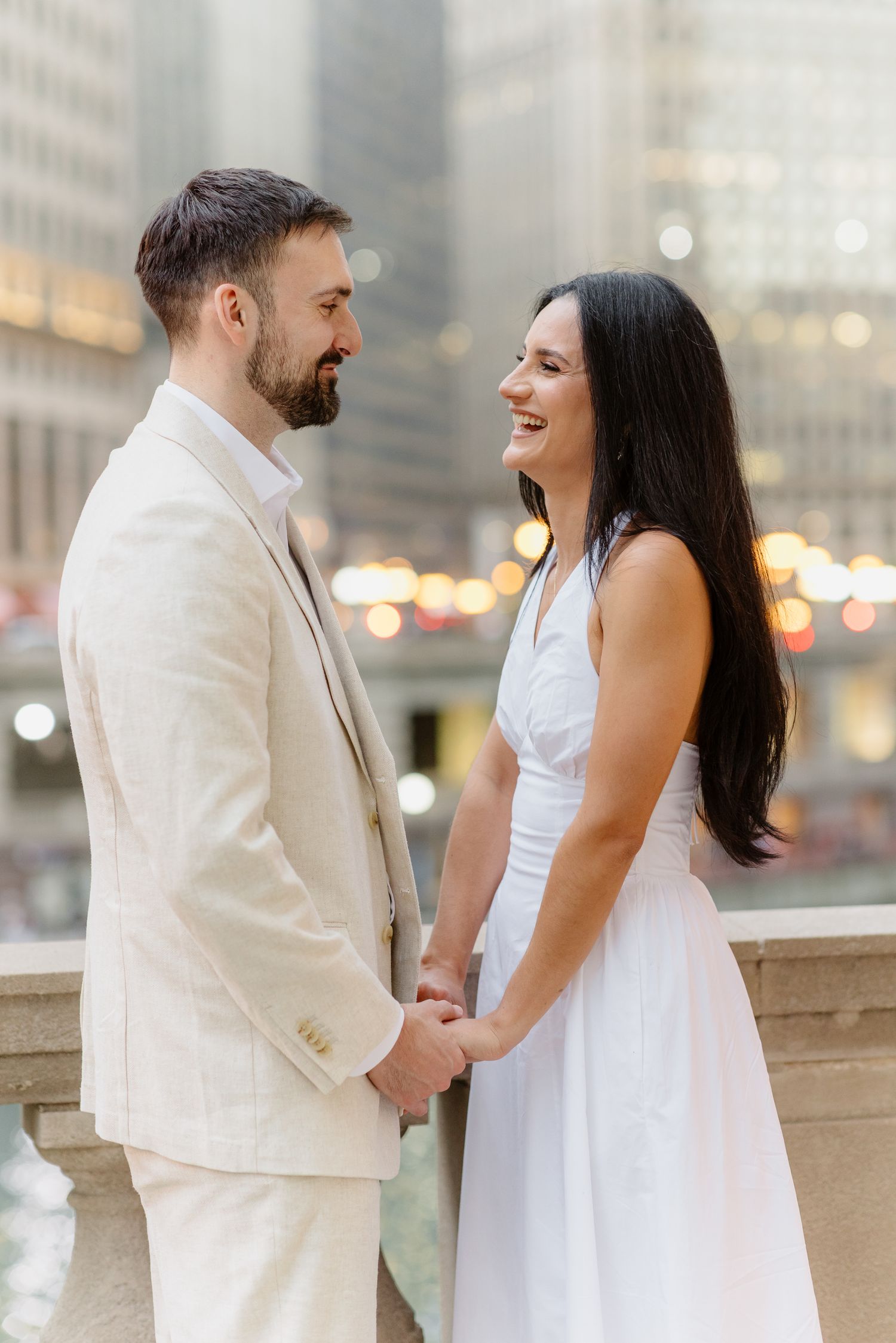 Happy couple in white holding hands and smiling at each other with urban bokeh backdrop.