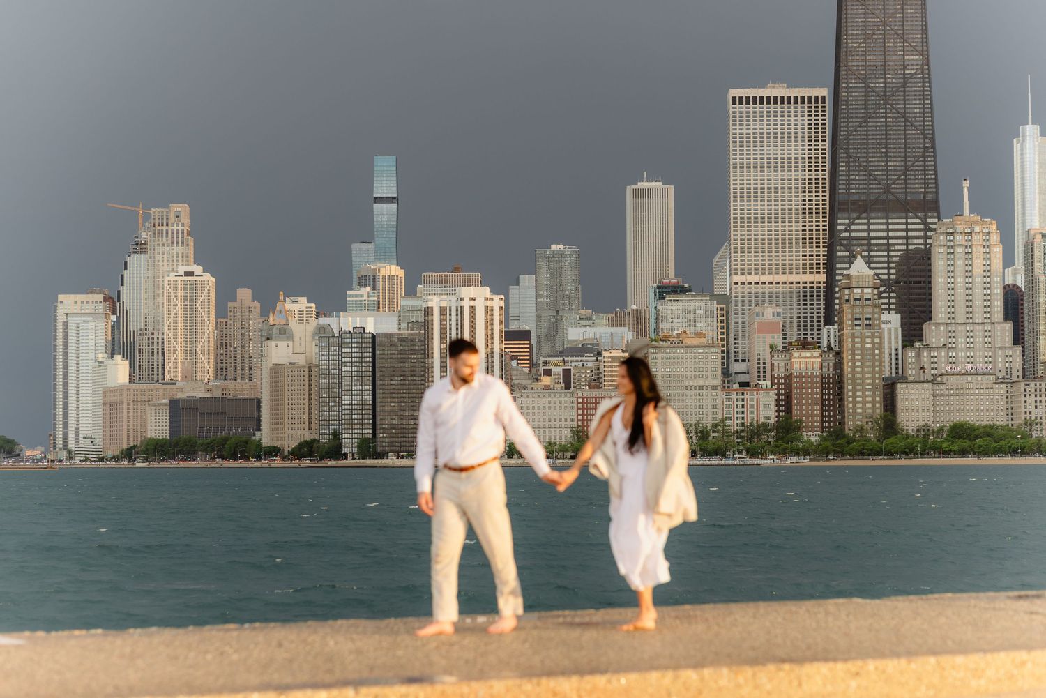 Engaged couple walking hand in hand along Chicago lakefront with dramatic skyline in the background.