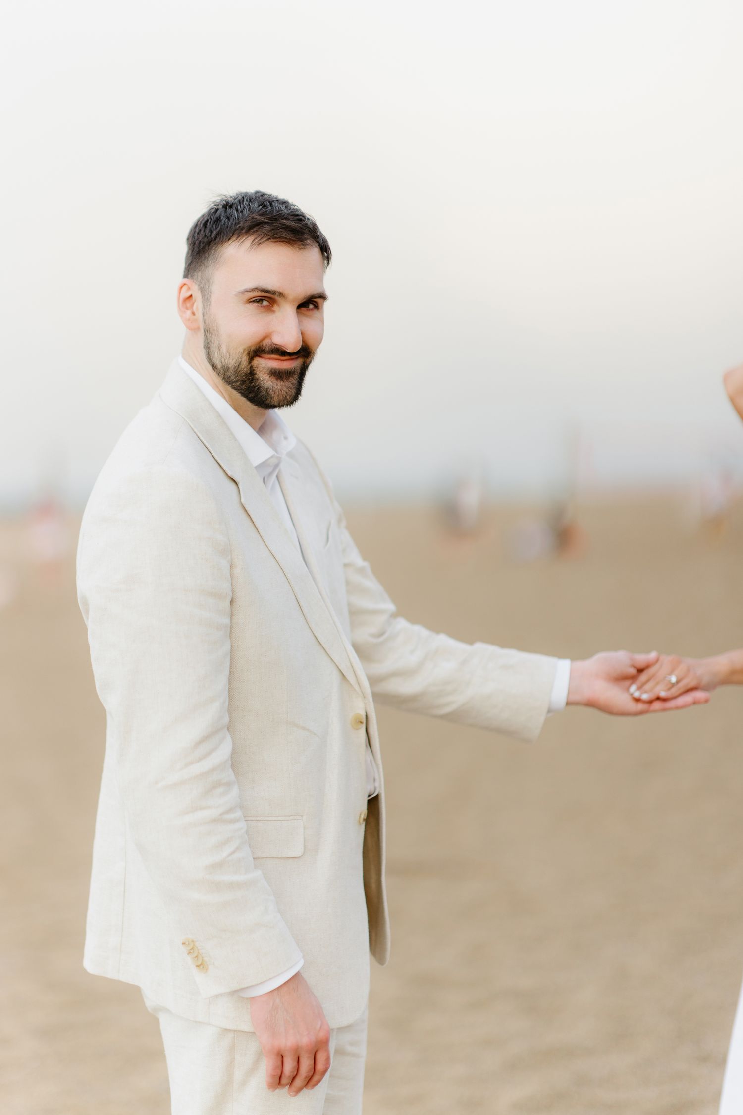 A bearded man in a beige suit smiles while holding hands with someone on a sandy beach.