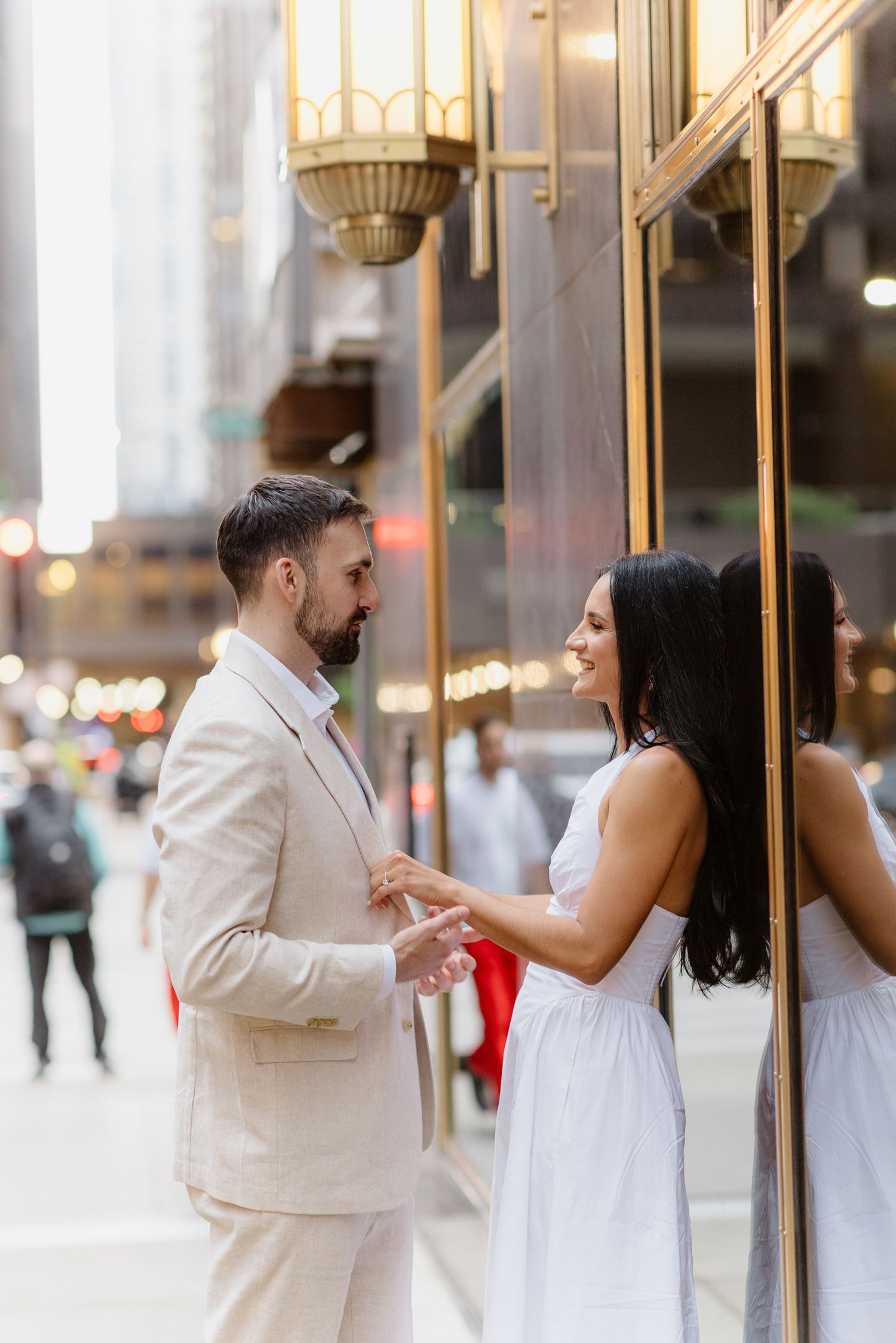 Engaged couple holding hands and gazing at each other beside gold-framed building on city street.